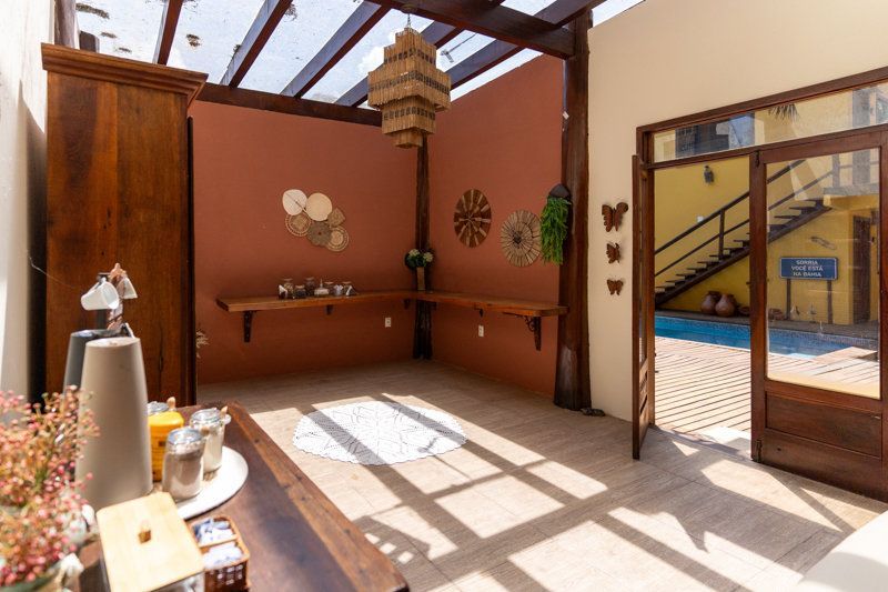 Pátio ensolarado com postes de madeira, paredes de terracota, plantas em vasos e uma porta de vidro aberta para um pátio interno.