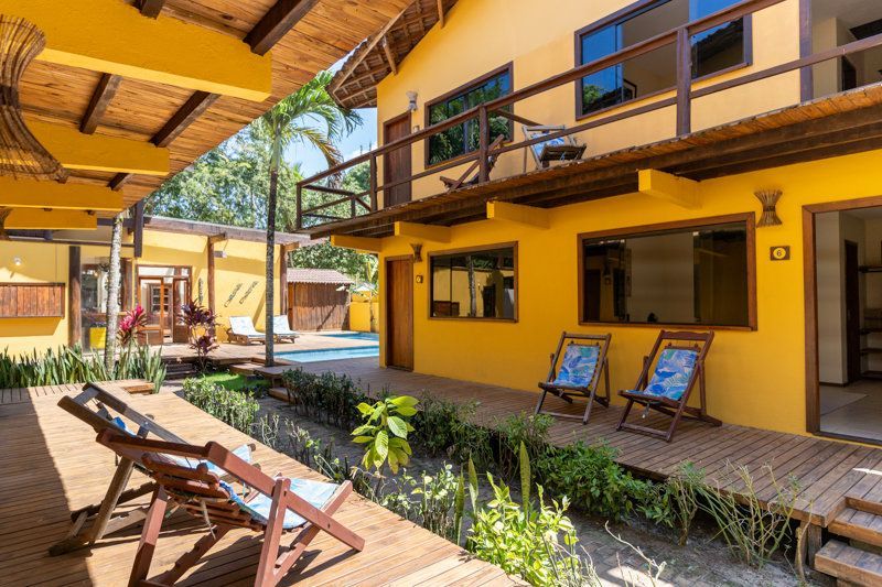 Pátio ensolarado de uma casa amarela de dois andares com espreguiçadeiras, varanda e uma pequena piscina.