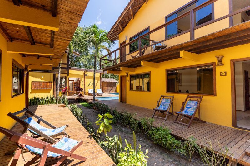 Pátio amarelo do hotel com espreguiçadeiras, plantas e uma passarela central entre as varandas.
