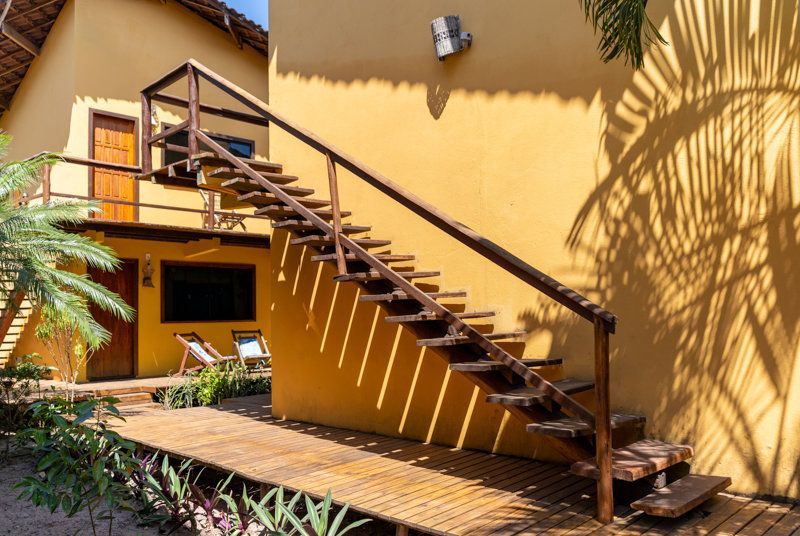 Escada externa de madeira com corrimão encostada em um prédio de estuque amarelo em um pátio ensolarado.