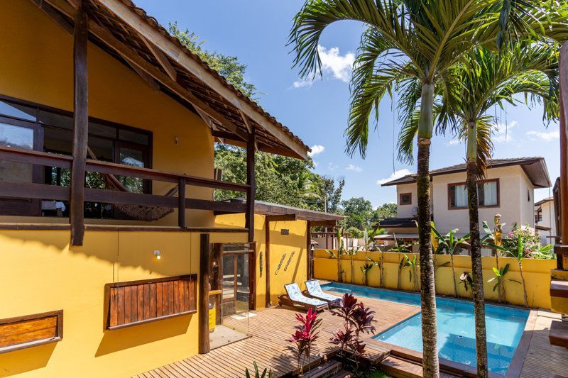 Pátio ensolarado de um resort tropical com prédios amarelos, palmeiras e uma pequena piscina azul.