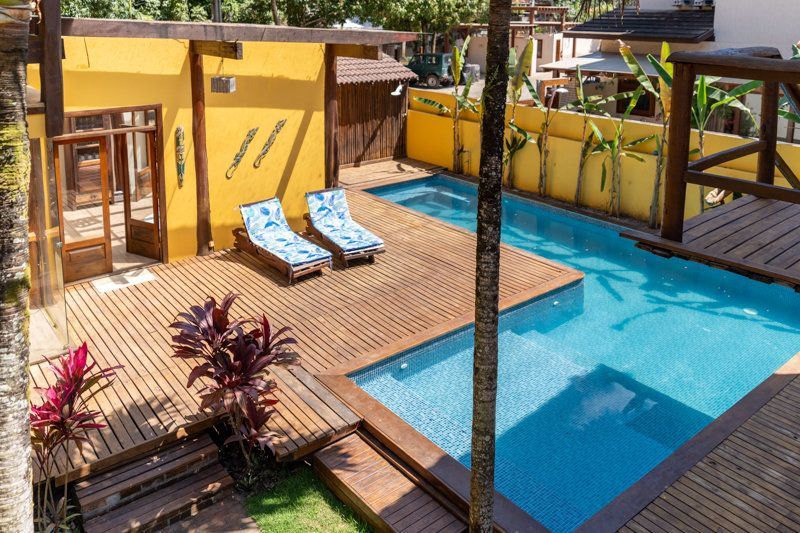 Pátio amarelo com piscina azul, duas espreguiçadeiras e vasos de plantas sob uma pérgola.