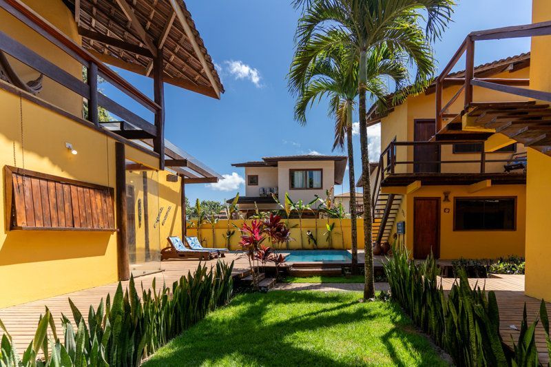 Pátio ensolarado de um resort tropical com edifícios amarelos, palmeiras e gramado verde ao redor de uma piscina central.