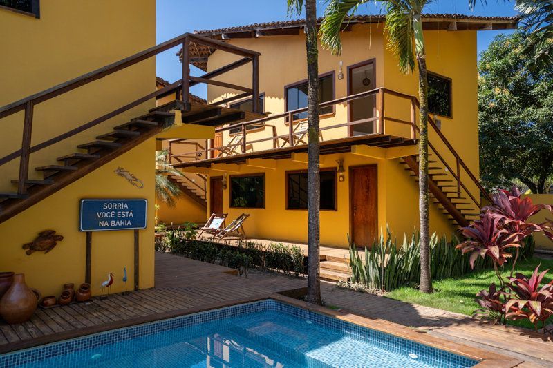 Prédio amarelo de um resort com escadarias de madeira ao lado de uma piscina azul e palmeiras.