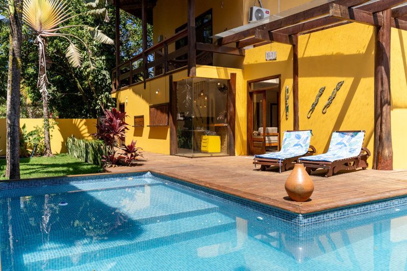 Pátio à beira da piscina com piscina turquesa, villa amarela e espreguiçadeiras em um cenário tropical.