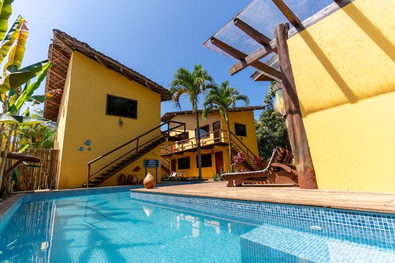 Pátio com piscina tropical iluminada, edifícios amarelos, palmeiras e céu azul.