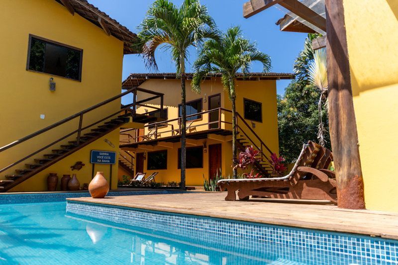 Pátio ensolarado com piscina turquesa, casa amarela e palmeiras.
