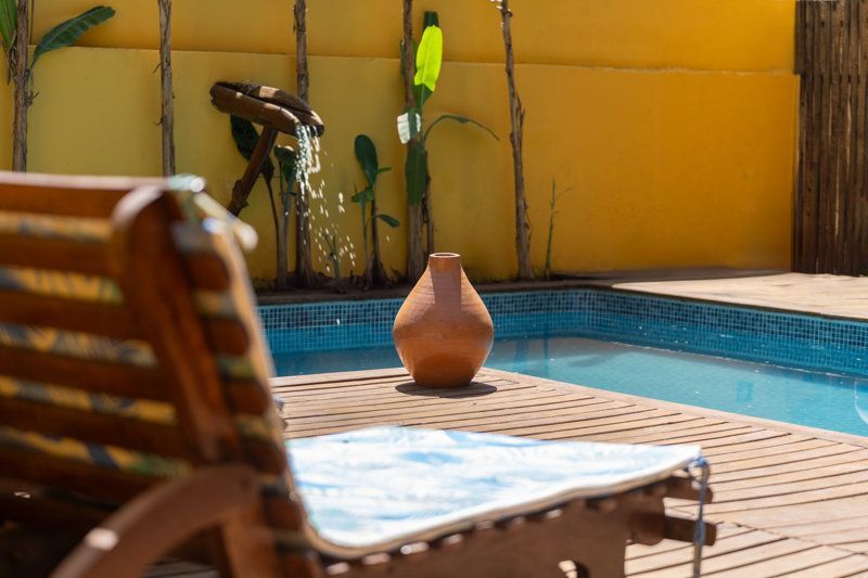 Pátio à beira da piscina com espreguiçadeira, vaso de barro e parede amarela junto à água azul.