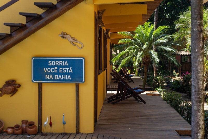 Blue sign reading “SORRIA VOCÊ ESTÁ NA BAHIA” beside a yellow house and wooden deck in a tropical garden