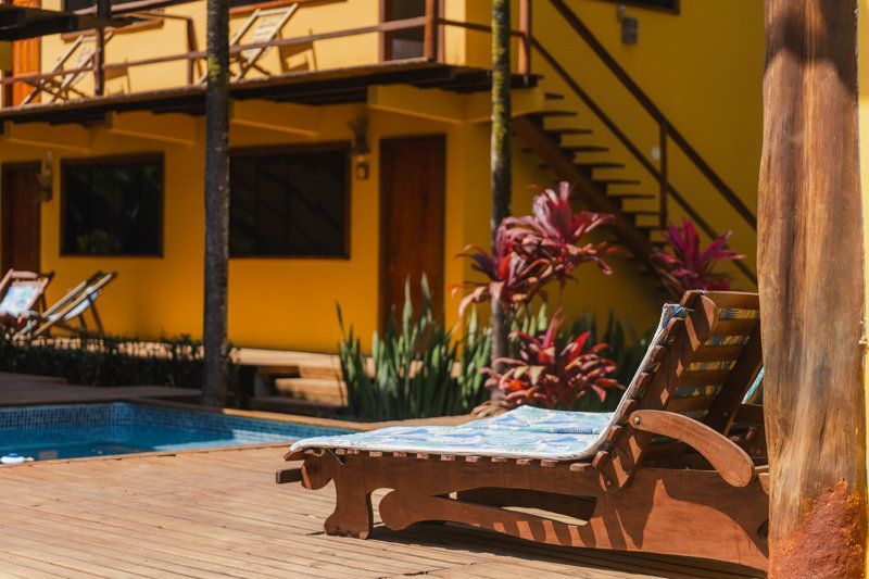 Pátio ensolarado à beira da piscina com espreguiçadeiras azuis, prédio amarelo e plantas tropicais.