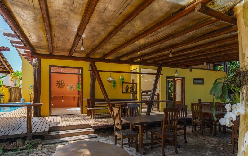 Pátio externo com mesas de madeira sob um telhado ripado, paredes amarelas e portas laranjas.