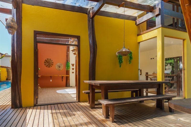 Pátio externo coberto com paredes amarelas, mesa e banco de madeira, e uma porta aberta para um cômodo interno laranja.
