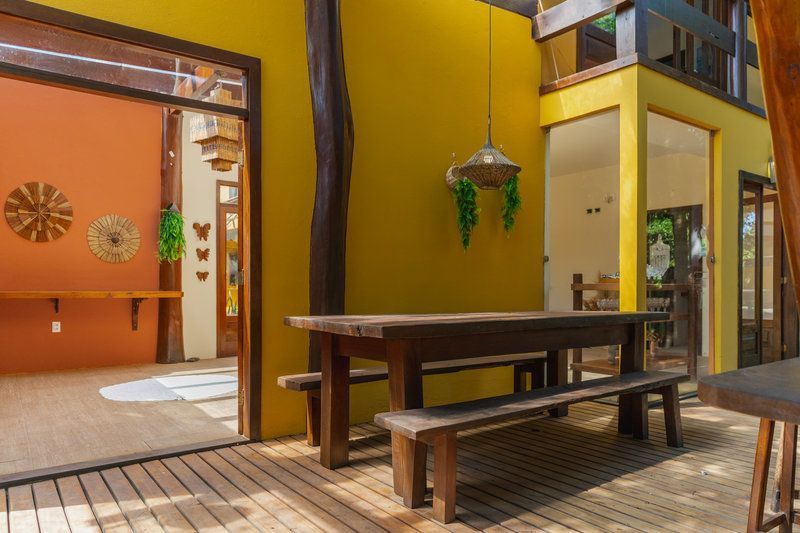 Pátio ao ar livre com mesa e bancos de madeira ao lado de um prédio amarelo e um cômodo interno laranja.