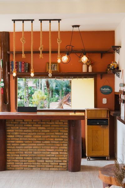 Interior de bar aconchegante com balcão de tijolos, luminárias pendentes, prateleiras de madeira e uma janela com vista para a vegetação.