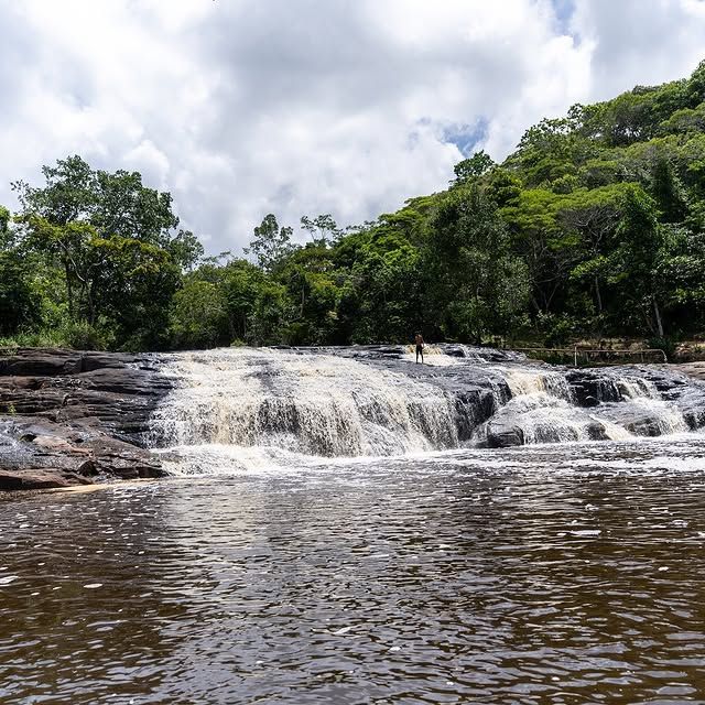 Pequena cachoeira que deságua em um rio marrom sobre rochas, cercada por uma densa floresta verde sob um céu nublado.