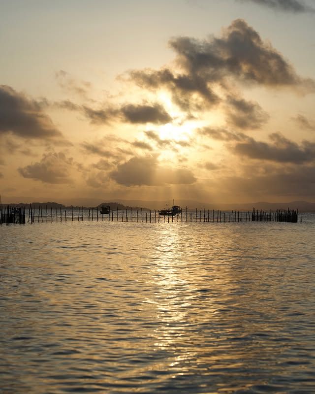 Pôr do sol sobre águas calmas com reflexos dourados e nuvens silhuetadas, com postes distantes no horizonte.