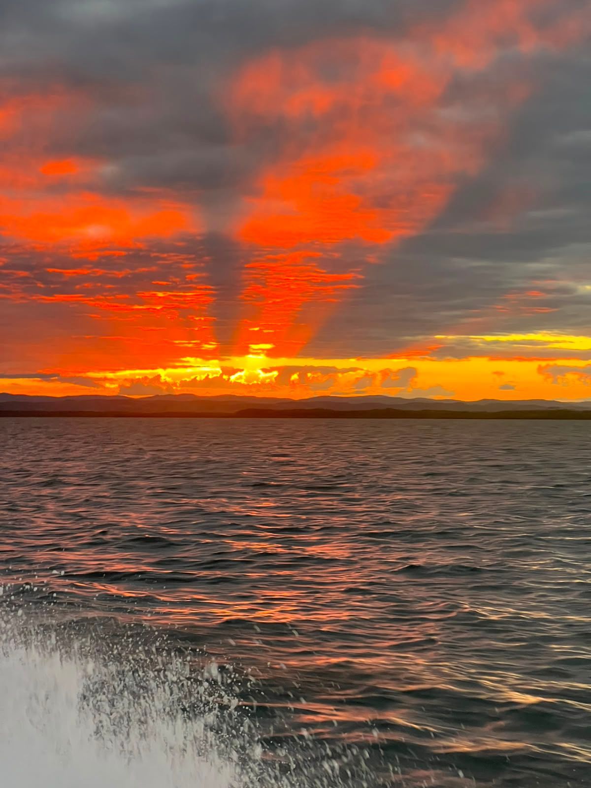Pôr do sol alaranjado sobre águas ondulantes com nuvens dramáticas e respingos em primeiro plano.