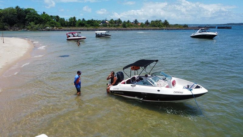 Lancha a uma praia rasa de areia com pessoas embarcando, vários barcos flutuando ao largo e uma costa arborizada.
