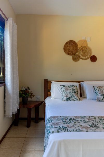 Quarto iluminado com cama branca, almofadas florais, decoração de parede, mesa de cabeceira e luz natural vinda da janela.