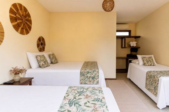Quarto bege claro com duas camas brancas, tapetes florais e decoração de parede em vime.