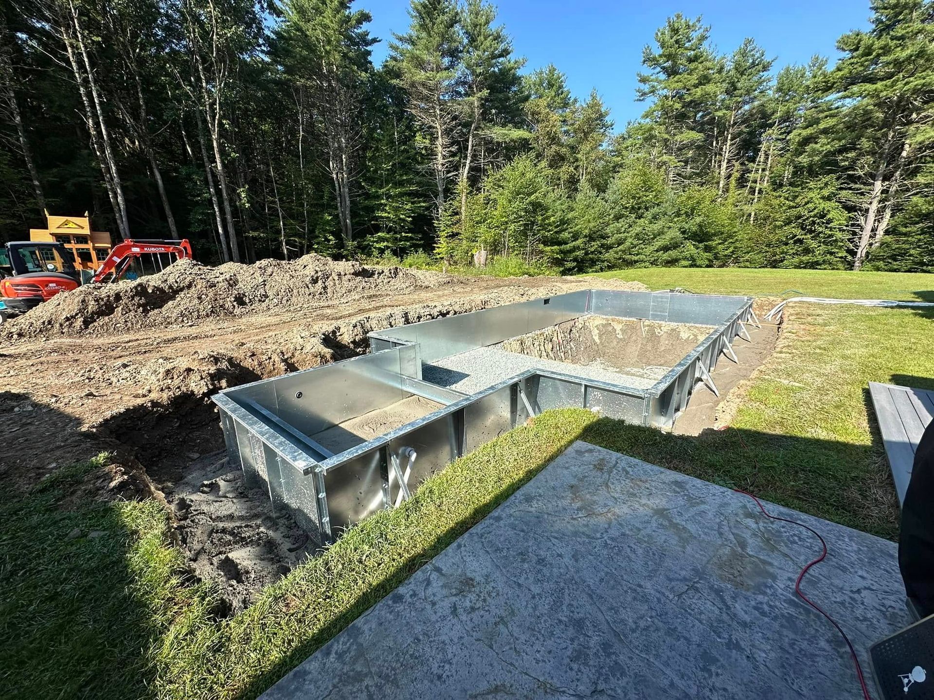 Pool construction site. Excavation of a rectangular and L-shaped structure in a grassy yard, with trees in the background.