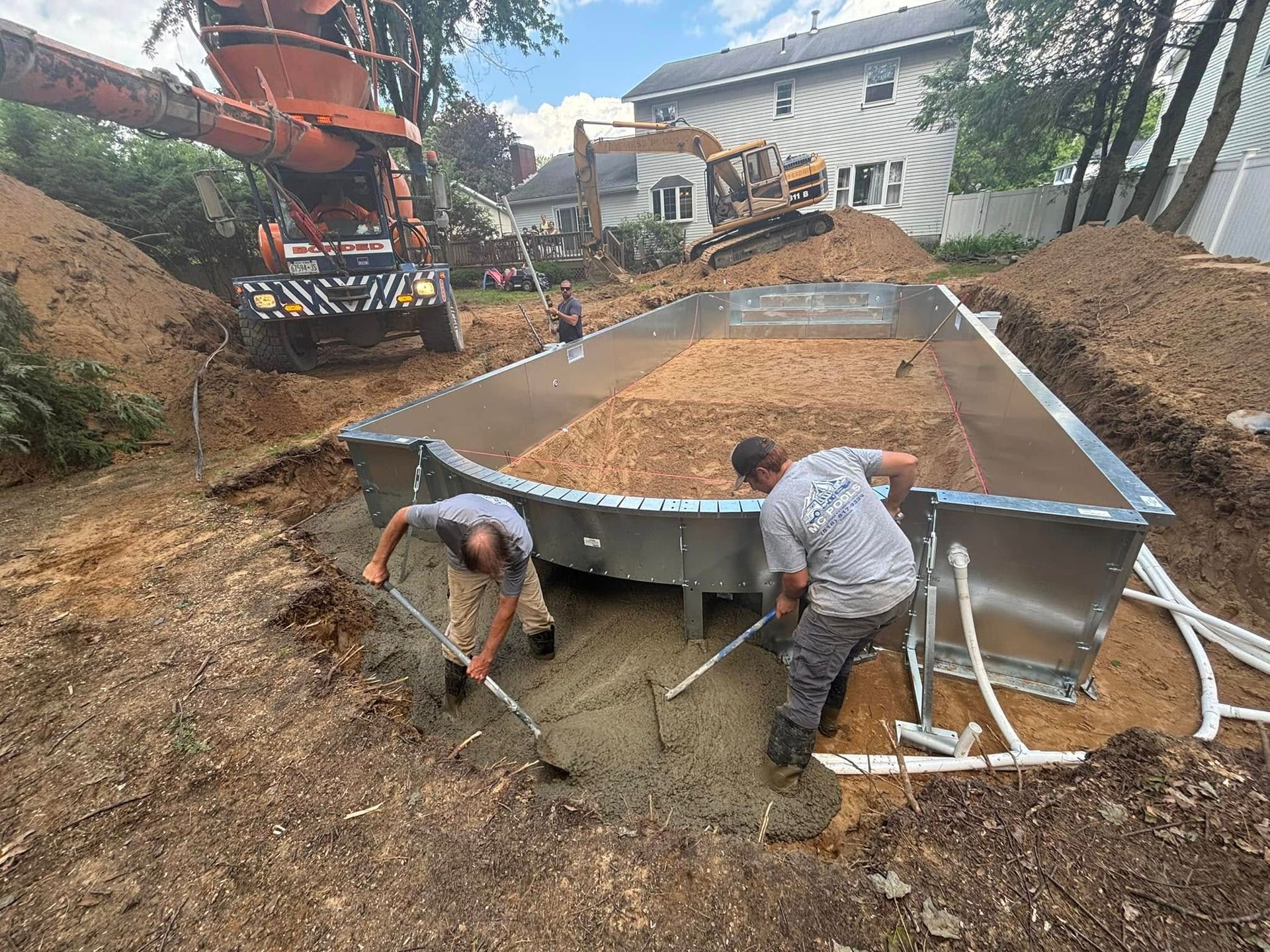 Two workers spreading concrete in a pool form. Construction equipment and a house are in the background.