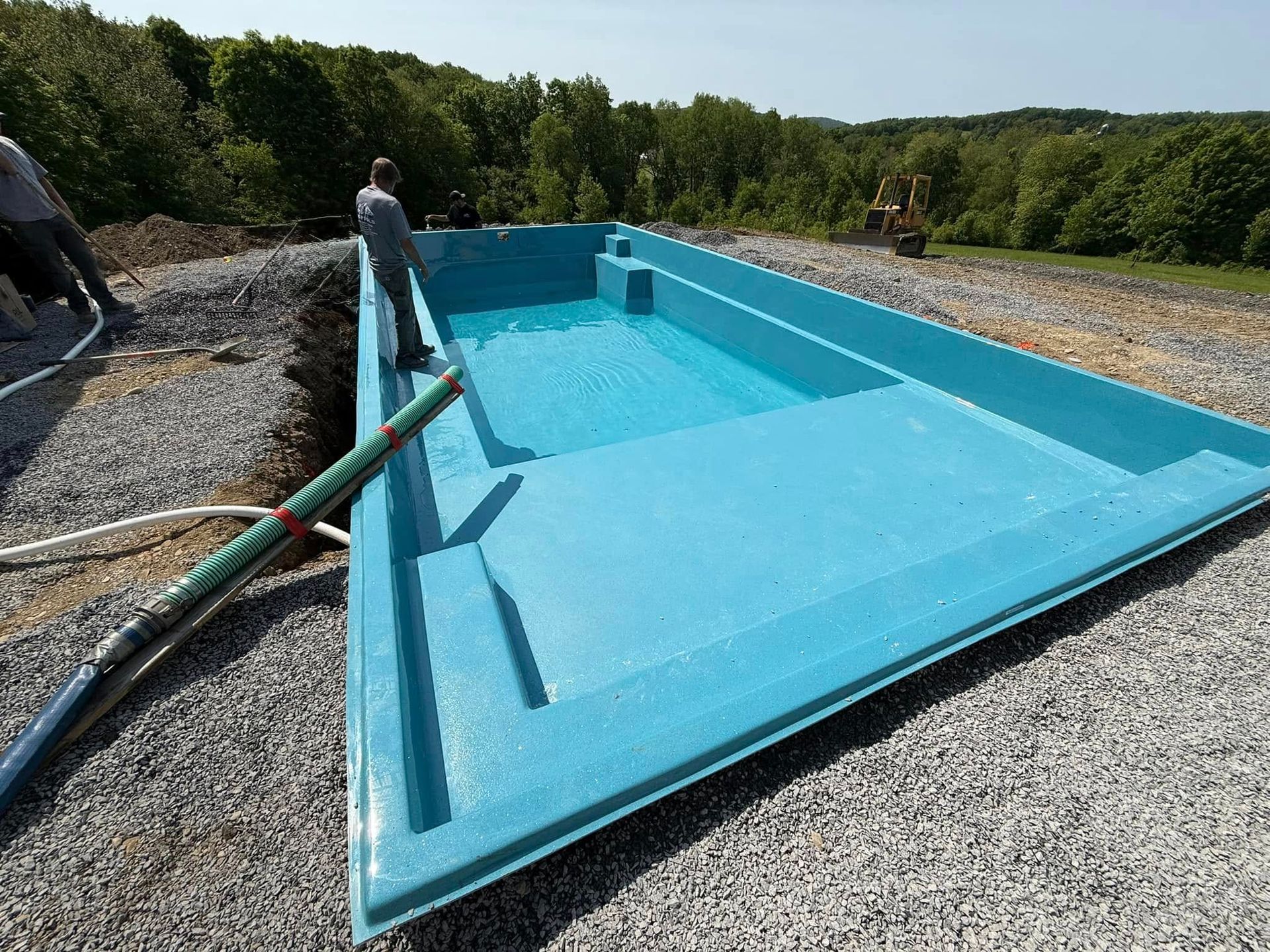 Blue fiberglass pool under construction, filled with water, surrounded by gravel, workers nearby.