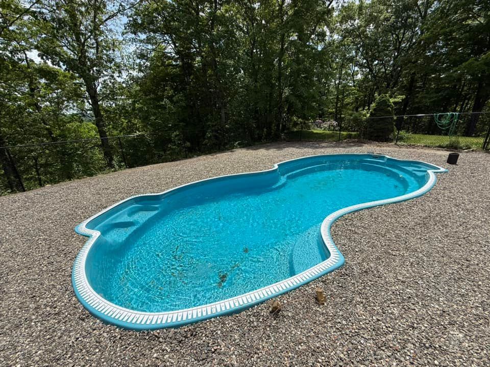 A blue, kidney-shaped swimming pool surrounded by gravel in a yard with trees.