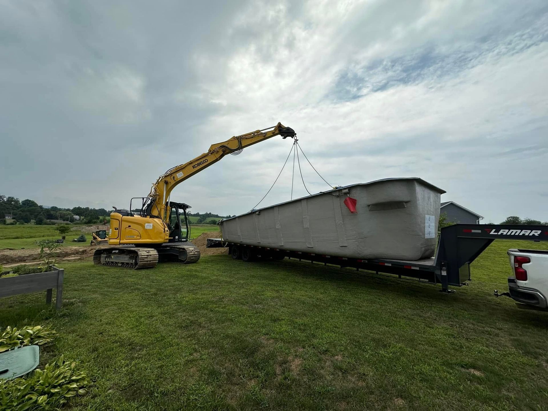 An excavator lifts a gray structure onto a trailer in a grassy field under a cloudy sky.