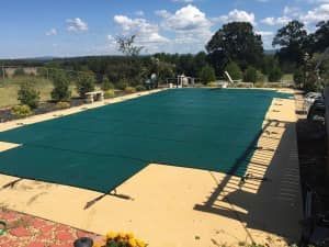 A green pool cover on a rectangular pool, surrounded by a light-colored patio, with trees and a blue sky in the background.