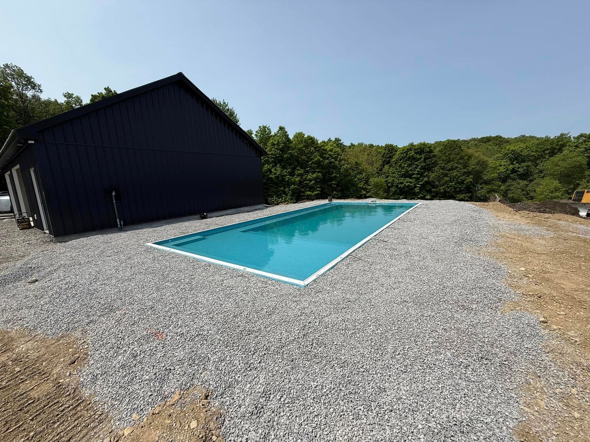 Rectangular pool filled with turquoise water next to a dark-sided building and surrounded by gravel, trees in the background.
