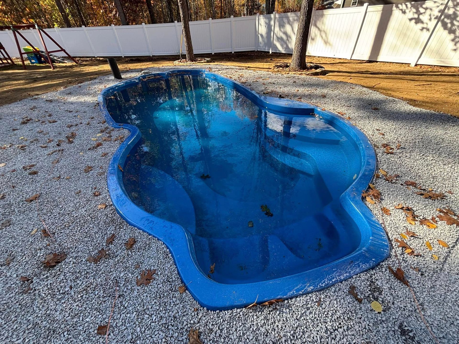 Blue, kidney-shaped inground pool filled with water, surrounded by gravel, trees, and a white fence.