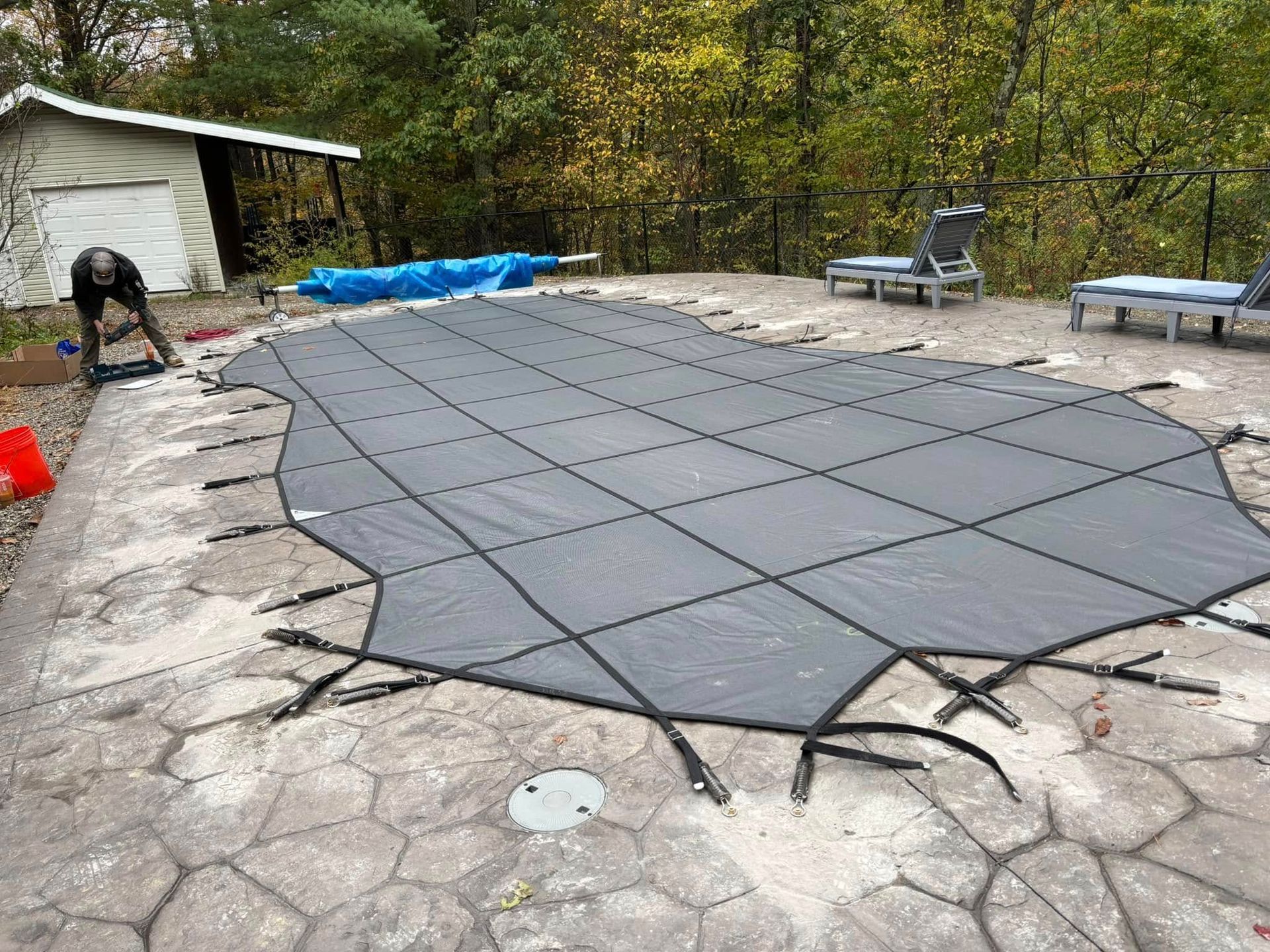 Pool covered with gray safety cover. A person is working near the pool. Trees in background.