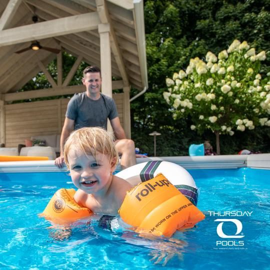 Child in orange floaties in a pool, smiling with an adult sitting on the deck behind.