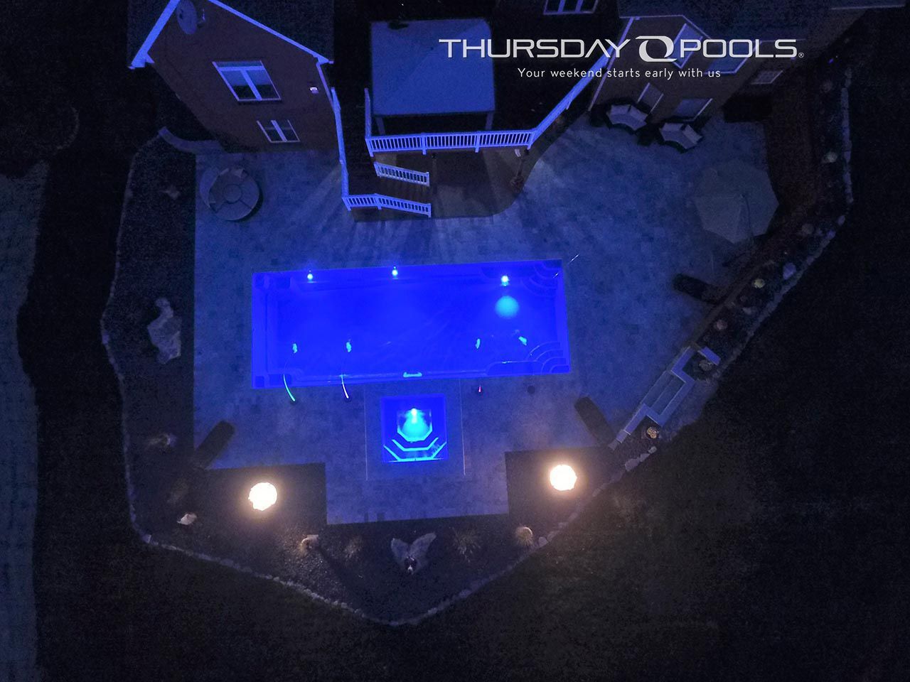 Blue-lit backyard pool deck at night with three bright lights and a house in the background
