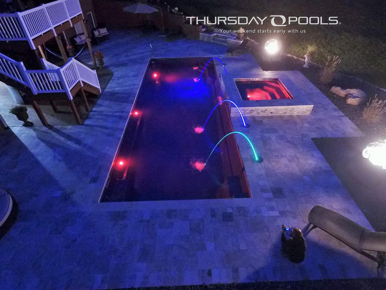 Nighttime backyard with a lit rectangular pool, hot tub, and lounge chairs on a stone patio