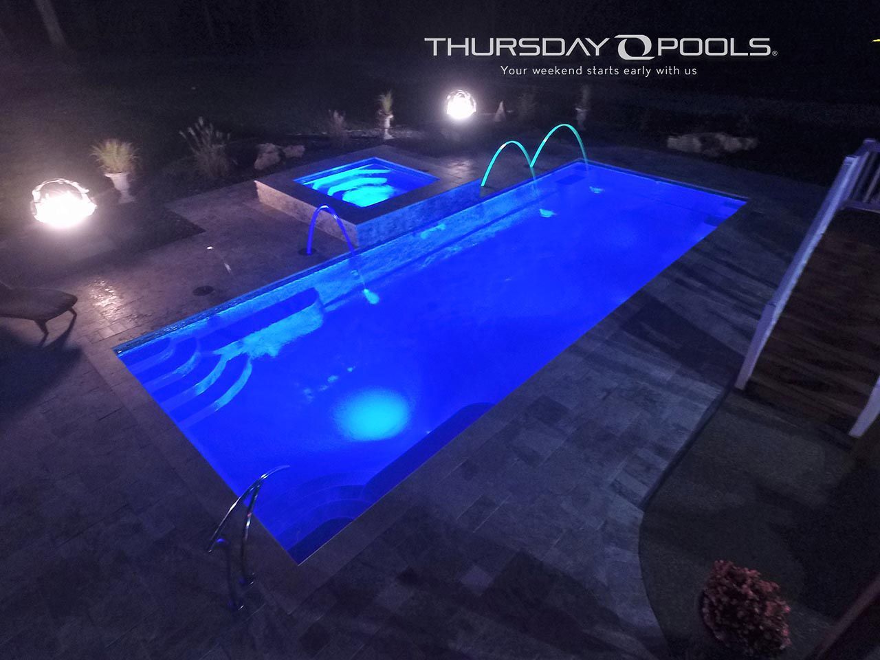Nighttime backyard pool glowing blue, with lit steps, fountain jets, and patio seating