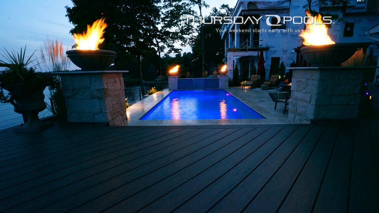 Blue-lit backyard pool with fire bowls and wooden deck at dusk