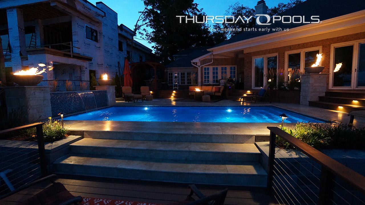 Lit outdoor pool at dusk with blue lights, steps, lounge chairs, and surrounding buildings.