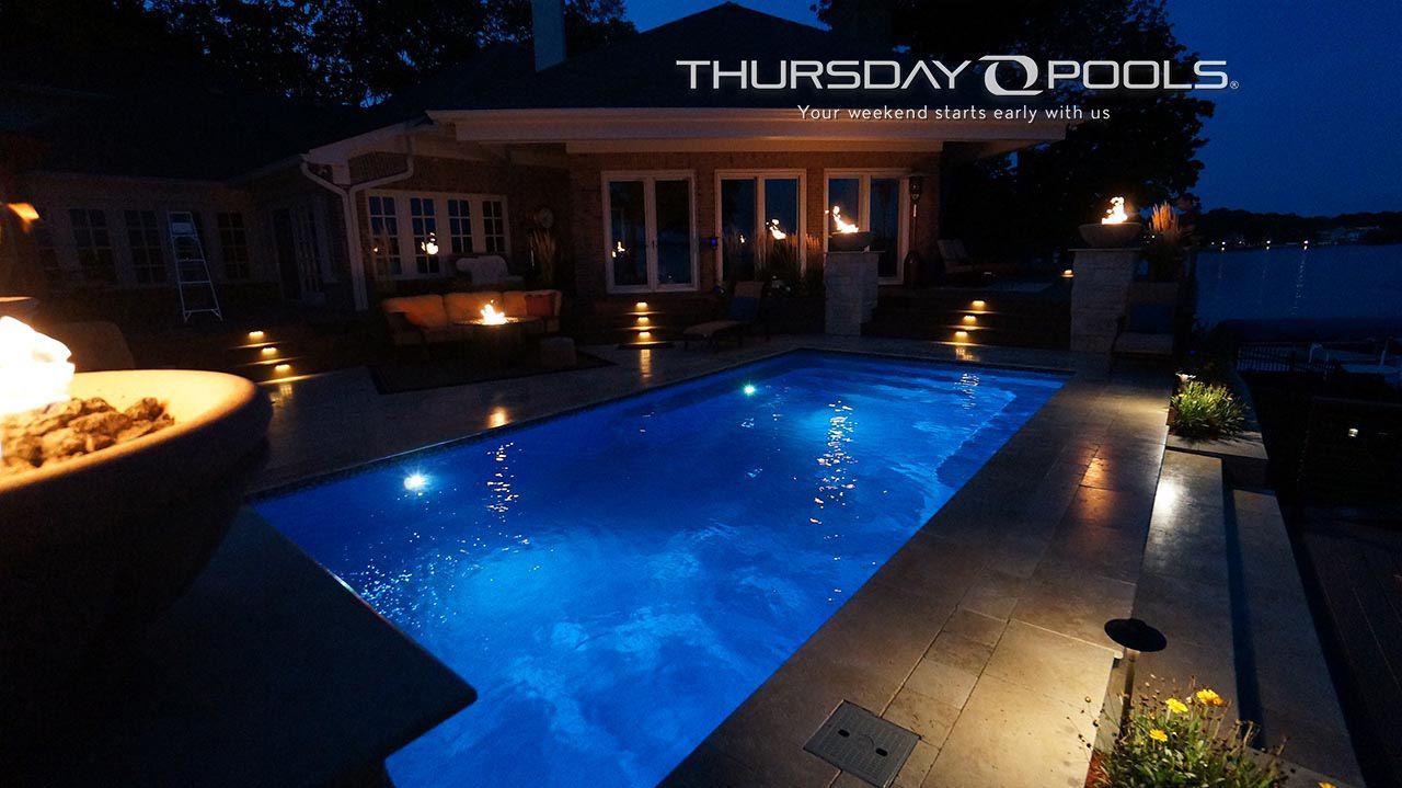 Nighttime view of a lit backyard pool with blue water and patio lights, under a Thursday Pools sign