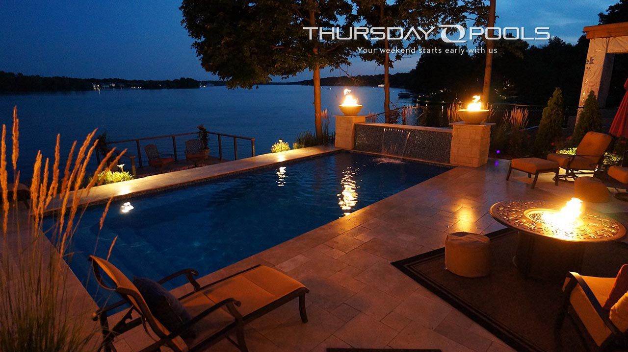 Poolside patio at dusk with lit fire pit, lounge chairs, and a calm lake view