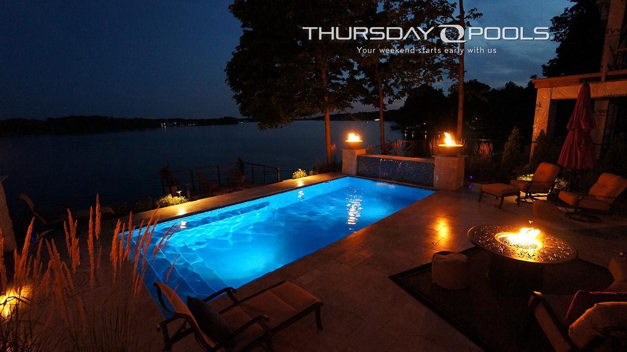 Illuminated blue swimming pool at dusk beside patio chairs and a fire pit, overlooking water and trees.