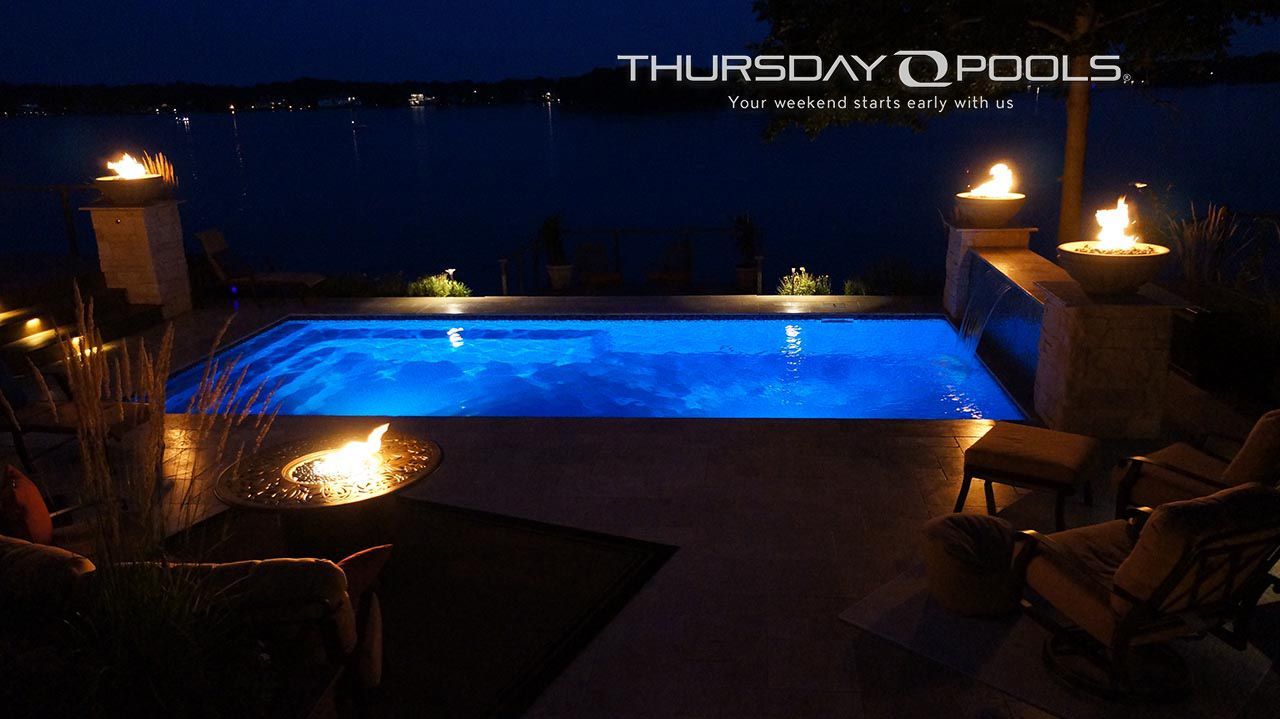 Nighttime backyard pool glowing blue, lit by torches and lanterns with lounge chairs around it