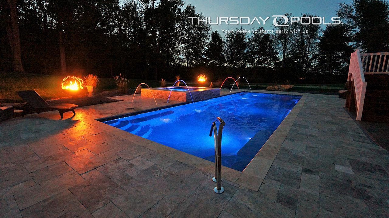 Illuminated backyard pool at night with blue water, stone patio, and surrounding fire lights