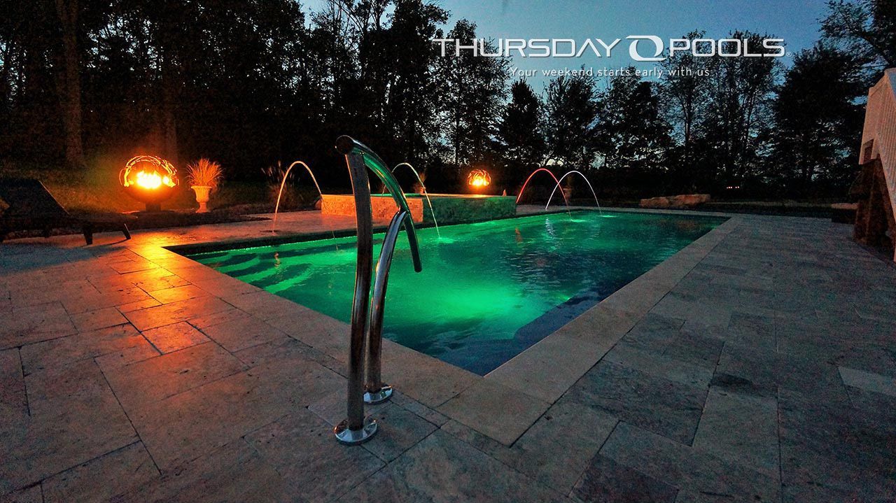 Lit swimming pool at dusk with green underwater lights and a pool ladder in the foreground