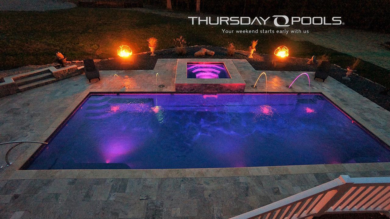 Lit backyard pool at dusk with purple underwater lights and fire bowls; “Thursday Pools” sign above.