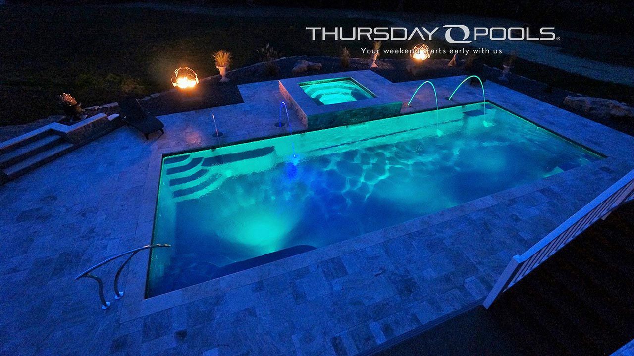 Nighttime backyard pool glowing blue with a hot tub and “Thursday Pools” sign