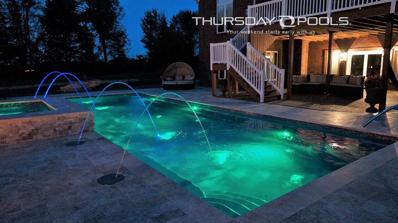 Backyard pool at dusk with glowing blue-green lights, fountain jets, and a lit patio and stairs nearby