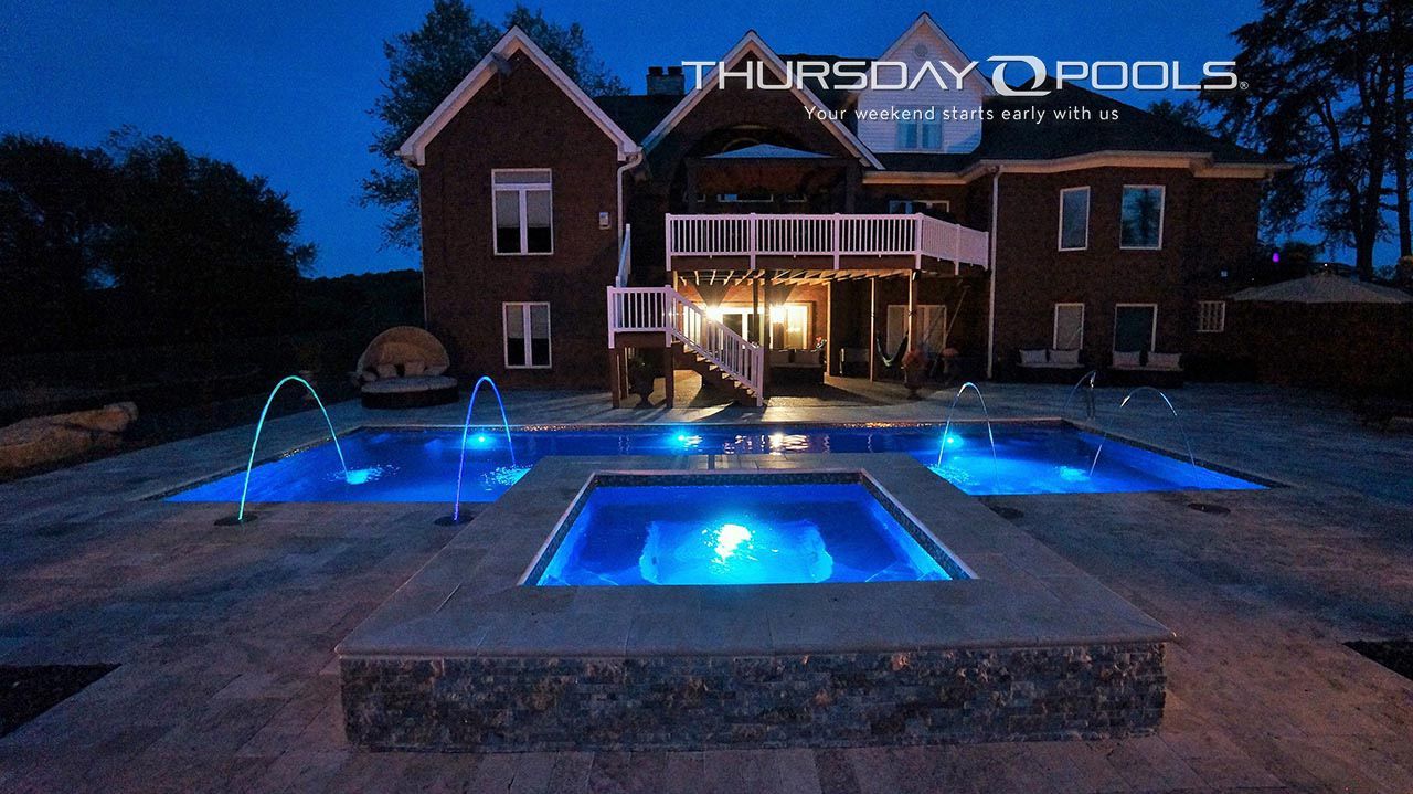 Blue-lit backyard pool and patio at dusk in front of a large brick house