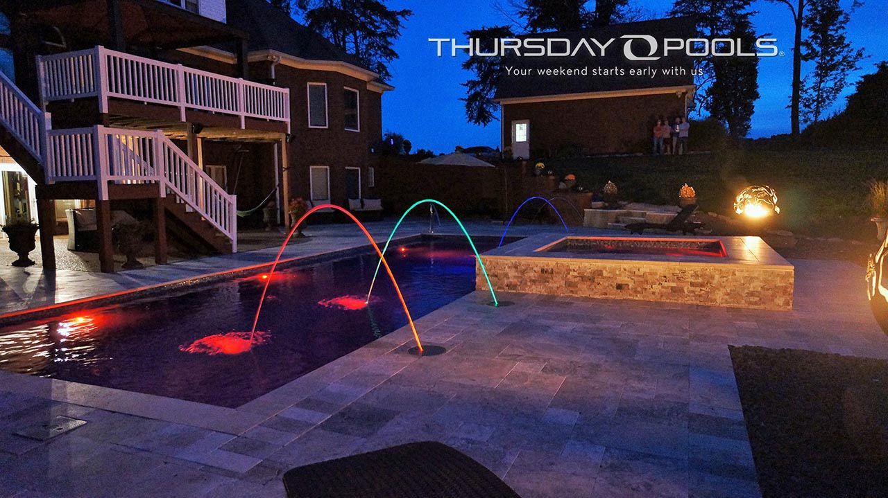 Backyard pool at dusk with lit water arcs and a house deck in the background